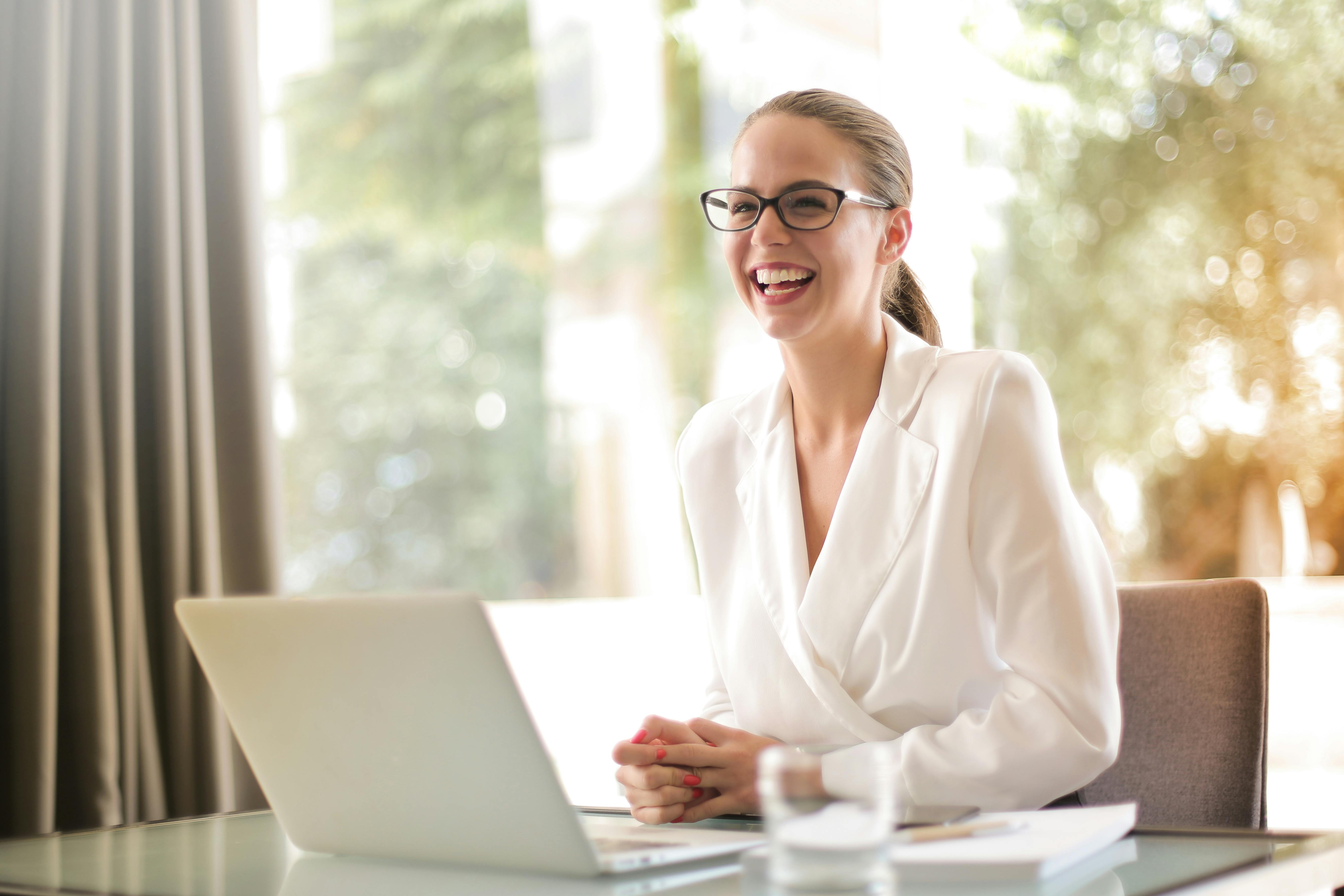 Career woman sitting and laughing with hands clasped on desk in front of laptop.